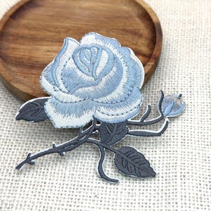 May include: Embroidered blue rose patch with gray stem and leaves. The flower has a light blue and white color scheme. The patch is on a textured, off-white surface, with a wooden plate in the background.