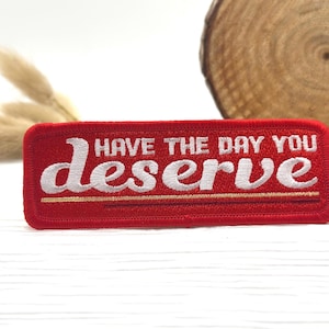 May include: A rectangular red patch with white embroidered text that reads "HAVE THE DAY YOU deserve". The patch has a gold border and a gold line beneath the word "deserve". The patch is on a white surface.