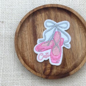 Peut inclure: Un écusson de chaussons de ballet roses avec des accents argentés et le mot "Cute" en cursive. L'écusson est sur un fond blanc avec un nœud argenté et est placé sur une assiette en bois ronde.