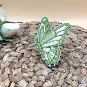 May include: Embroidered butterfly patch in shades of green and white. The butterfly is detailed with stitched lines, and the wings are outlined in white. The patch is placed on a woven, textured surface, with a white rose and green leaves in the background.