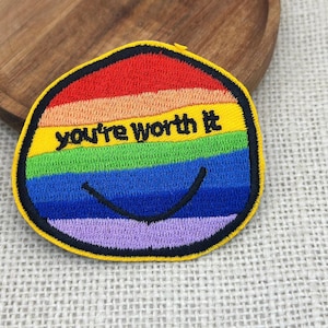 May include: Embroidered patch with a rainbow design and the words "you're worth it" in black. The patch has a yellow border and a smiling face. The patch is on a wooden surface.