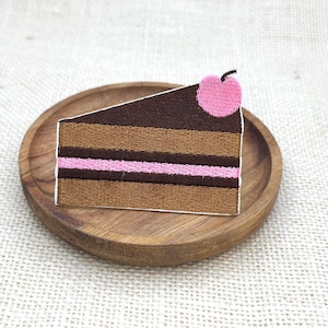 Op de afbeelding: Een driehoekige taartpunt-patch met donkerbruine, lichtbruine en roze horizontale lagen en een kleine roze kers bovenop. De geborduurde patch heeft de vorm van een stuk taart met een witte rand.