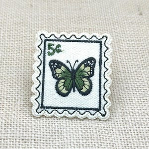 Puede incluir: Un parche bordado rectangular con forma de sello postal y bordes festoneados. El parche presenta un fondo blanco, un borde negro y una mariposa verde en el centro. El texto 5¢ está bordado en color verde en la esquina superior izquierda.