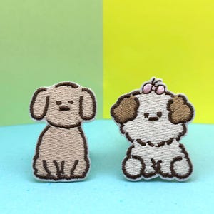 May include: Two embroidered iron-on patches of dogs. One is a brown and white poodle with brown ears and a white body. The other is a white poodle with brown ears and a pink bow.
