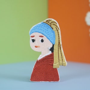 Puede incluir: Parche bordado de una niña con una diadema azul y un pendiente de perla, que lleva un vestido rojo y una larga trenza rubia.