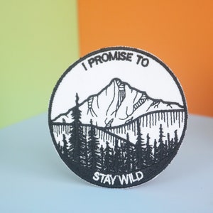 May include: A white embroidered patch with a black mountain range and trees. The text "I Promise to Stay Wild" is embroidered in black around the edge of the patch.