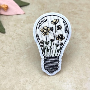 May include: Embroidered patch in the shape of a lightbulb, featuring a floral design. The lightbulb is white with a black outline, and the flowers are yellow and green. The base of the lightbulb is gray.