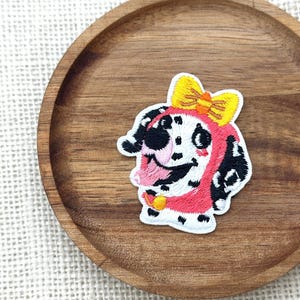 May include: Embroidered patch featuring a cartoon Dalmatian dog with a pink hood and a yellow bow. The dog has black spots and a smiling expression. The patch is on a wooden tray.