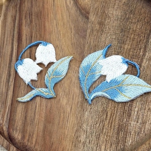 Può includere: Due toppe ricamate raffiguranti fiori di mughetto. Ogni toppa presenta fiori bianchi a forma di campana con steli e foglie azzurre, arricchiti da sottili dettagli in filo metallico dorato sui motivi floreali e sulle foglie.