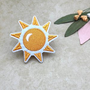 May include: Embroidered sun patch with a yellow center and white and yellow rays. The sun has a white crescent detail. The patch is on a neutral background with decorative leaves and flowers.