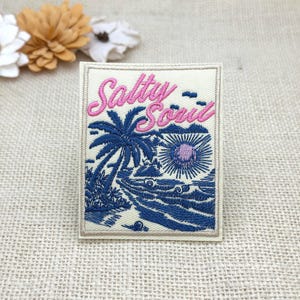 May include: A rectangular embroidered patch featuring a cream background with the pink text Salty Soul. The design depicts a dark blue palm tree, a sun with a purple centre, and ocean waves. The patch has a stitched border and a textured finish.