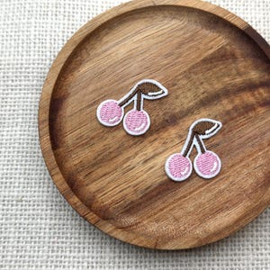 May include: Two embroidered cherry patches with pink fruit and brown stems, outlined in white. The patches are on a round, wooden tray with a natural wood grain. The tray sits on a textured, off-white surface.