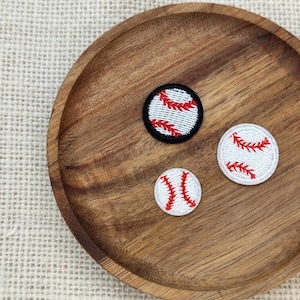 Può includere: Tre toppe da baseball di diverse dimensioni sono appoggiate su un piatto di legno. Le toppe presentano uno sfondo bianco con cuciture rosse, che imitano una palla da baseball. Una toppa ha un bordo nero.