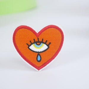 May include: Orange heart-shaped embroidered patch with a white eye and a blue teardrop. The eye has black eyelashes and a yellow pupil.