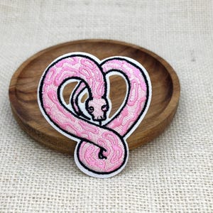 May include: An embroidered patch featuring a light pink snake with darker pink patterned markings, coiled into a heart shape. The snake has a black outline, a small head with visible eyes, and a white border surrounding the entire design.