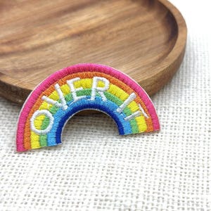Pode incluir: Patch de arco-íris bordado com a frase "OVER IT" em letras brancas. O patch apresenta uma gama vibrante de cores, incluindo rosa, laranja, amarelo, verde, azul e roxo. Um acessório divertido para roupas ou bolsas.