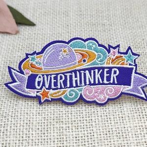May include: Embroidered patch with the word "OVERTHINKER" in purple script on a banner. The patch features a purple planet, stars, and colorful swirls in orange, teal, and pink. The patch is on a textured, neutral background.