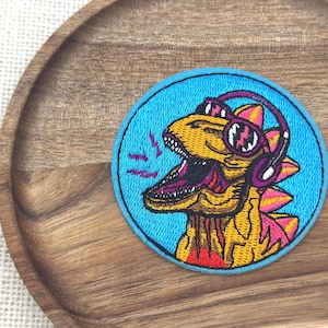 Può includere: Toppa ricamata raffigurante un dinosauro giallo con cuffie e occhiali da sole, su sfondo blu. Il dinosauro ha la bocca aperta con la lingua e i denti rosa. La toppa è rotonda con bordo nero.