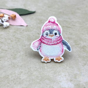 Peut inclure: Écusson de pingouin brodé portant un bonnet et une écharpe roses. Le pingouin a un ventre blanc, des ailes grises et des pieds orange. L'écusson est sur un fond neutre avec quelques feuilles et fleurs décoratives.