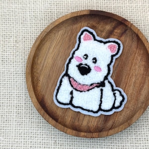 May include: A white, fluffy dog patch with a pink scarf and blush. The patch is outlined in black and sits on a wooden plate. The dog has a black nose and small black eyes. The background is a light tan burlap.