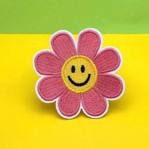 May include: Embroidered flower patch with a pink daisy design, a yellow smiley face center, and a white border. The patch is on a yellow and green background. This patch can be ironed on or sewn.