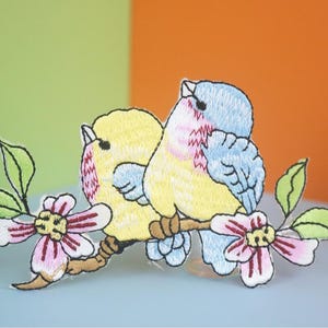 May include: Embroidered patch featuring two yellow and blue birds perched on a branch with pink flowers. The birds are facing each other and have detailed embroidery.