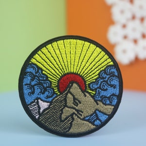 May include: A round embroidered patch with a mountain scene. The mountain is brown and has a red sun setting behind it. The sky is blue with yellow rays of light coming from the sun.