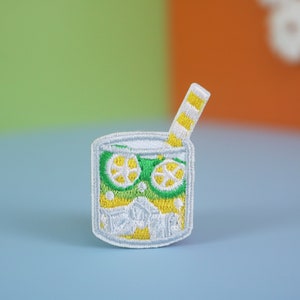 May include: White embroidered patch of a glass of lemonade with a yellow and white striped straw. The glass is filled with green and yellow lemon slices and white ice cubes.