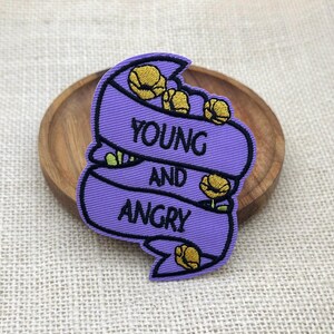 May include: A purple embroidered patch shaped like a ribbon banner with black stitched borders. The text "YOUNG AND ANGRY" is embroidered in black capital letters across the center. Small yellow flower buds are scattered throughout the design.