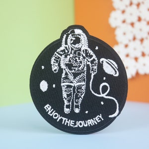 May include: Black embroidered patch with a white astronaut in a space suit, a planet, and a star. The text "Enjoy the Journey" is written in a white arc around the astronaut.