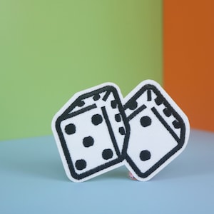 Peut inclure: Deux dés blancs avec des points noirs, cousus sur un patch de tissu blanc. Les dés se chevauchent partiellement, l'un faisant face à l'avant et l'autre sur le côté.