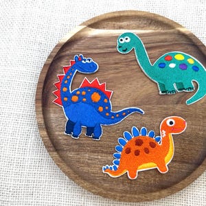 May include: Three colorful dinosaur patches on a wooden plate. The patches include a blue dinosaur with orange spots and a red spine, a green dinosaur with colorful spots, and an orange dinosaur with blue spines.
