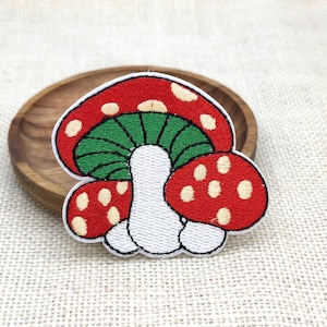 May include: Embroidered mushroom patch featuring a red cap with white spots, a green underside, and a white stem. The patch is outlined in black and sits on a wooden surface, perfect for adding to clothing or accessories.