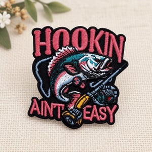 May include: An embroidered patch featuring a black background with a stylized fish and a fishing rod. The text "HOOKIN" is stitched in pink at the top, and "AINT EASY" is stitched in pink at the bottom. The fish is blue, white, and pink.