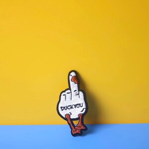 May include: A white embroidered patch of a duck giving the middle finger with the text "Duck You" on the palm. The patch is on a yellow and blue background.