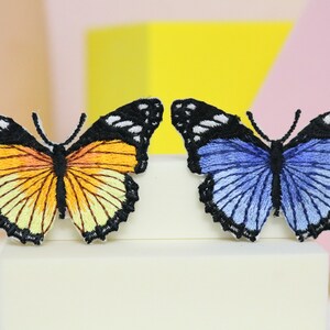 Puede incluir: Dos parches de mariposa bordados, uno amarillo y otro azul, con contornos negros y detalles blancos.
