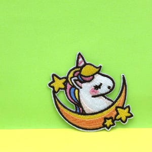 May include: A white unicorn with a rainbow mane and tail is sleeping on a crescent moon with stars. The unicorn has closed eyes and pink cheeks. The moon is orange and yellow.