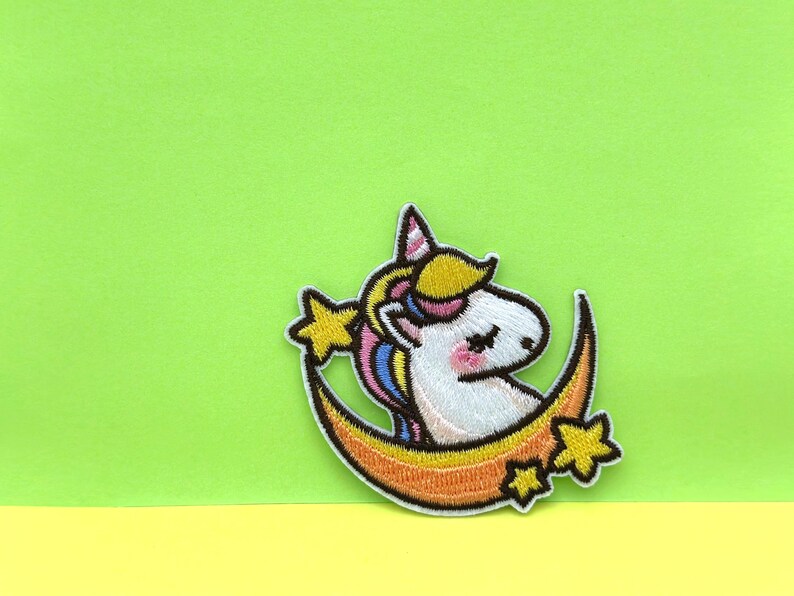 May include: A white unicorn with a rainbow mane and tail is sleeping on a crescent moon with stars. The unicorn has closed eyes and pink cheeks. The moon is orange and yellow.