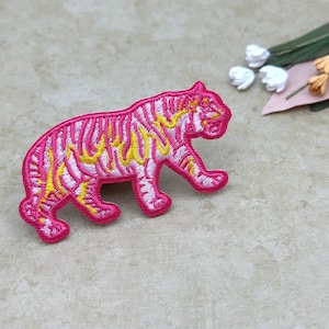 Puede incluir: Parche bordado de un tigre rosa con rayas amarillas y blancas. El tigre está representado caminando. El parche está sobre una superficie beige claro, con flores decorativas en el fondo. Ideal para personalizar ropa y accesorios. Mide aproximadamente 7 cm.