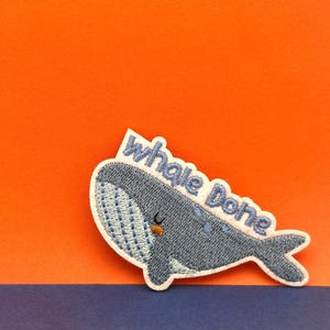 May include: A blue whale embroidered patch with the text "Whale Done" in white. The patch is on a red and blue background.