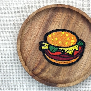 May include: Embroidered patch featuring a cartoon hamburger with a sesame seed bun, lettuce, tomato, and a burger patty. The patch has a black border and is set on a wooden plate.