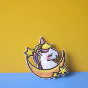 May include: A white unicorn with a rainbow mane and tail is sleeping on a yellow crescent moon with stars. The unicorn is embroidered on a white fabric patch.