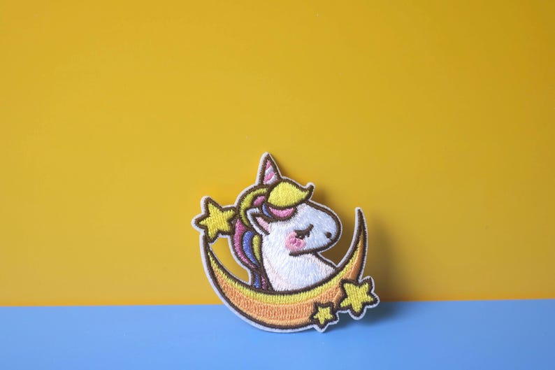 May include: A white unicorn with a rainbow mane and tail is sleeping on a yellow crescent moon with stars. The unicorn is embroidered on a white fabric patch.