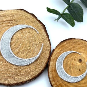 May include: Two silver crescent moon patches on wooden slices. The patches have a textured, embroidered appearance. The wood slices have a natural, rustic look with visible grain and bark. A sprig of green leaves is visible in the background.