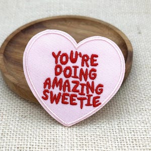 May include: A heart-shaped embroidered patch in light pink with red stitched text that reads YOU'RE DOING AMAZING SWEETIE. The patch features a clean, finished border and is designed for application onto fabric surfaces as a decorative accent.