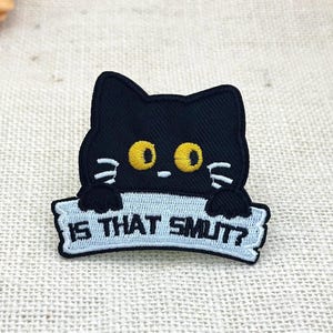 Puede incluir: Un parche bordado que muestra un gato negro con grandes ojos amarillos y bigotes blancos. El gato sostiene una pancarta azul claro con el texto negro IS THAT SMUT? El parche tiene un borde negro y tiene forma de cabeza y patas de gato.