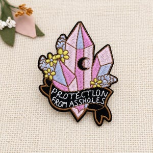 May include: An embroidered patch featuring pink and blue crystals, yellow flowers, and a black crescent moon. A black banner at the bottom contains the white embroidered text "PROTECTION FROM ASSHOLES" with a brown border outline.