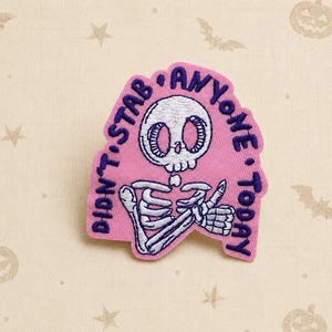May include: A pink embroidered patch featuring a white skeleton with large eyes and a small mouth. The skeleton is giving a thumbs up. The text DIDN'T STAB ANYONE TODAY is embroidered in dark purple thread in an arc around the skeleton figure.