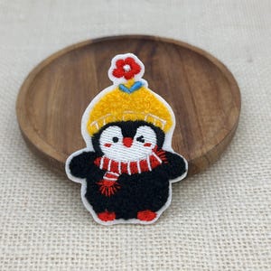 以下が含まれることがあります： 白いお腹をした黒いペンギンの刺繍ワッペン。黄色いニット帽の頂点には小さな赤い花があしらわれ、赤と白のストライプのマフラーを巻いています。ペンギンはウィンクをしており、赤い頬と赤い足が特徴です。