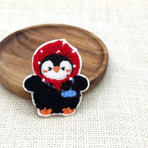 May include: Embroidered penguin patch. The penguin is wearing a red hooded jacket and carrying a blue bag. It has a black body, white face, and orange feet and beak. The patch is on a wooden plate.